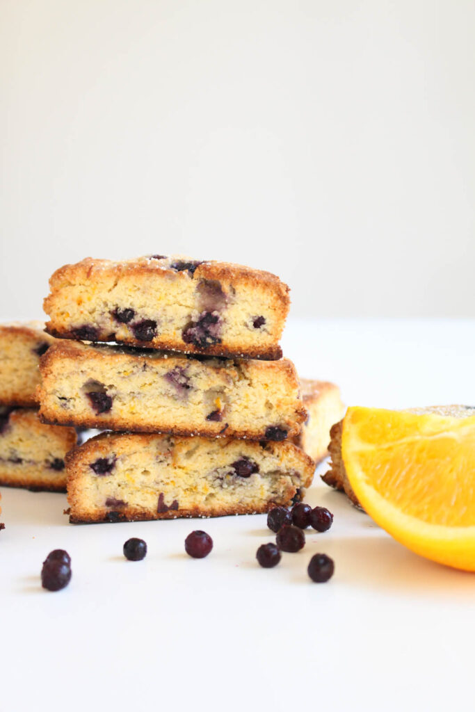 paleo blueberry scones stacked on top of each other, on a white background. Orange slices and blueberries for garnish in the front for garnish.