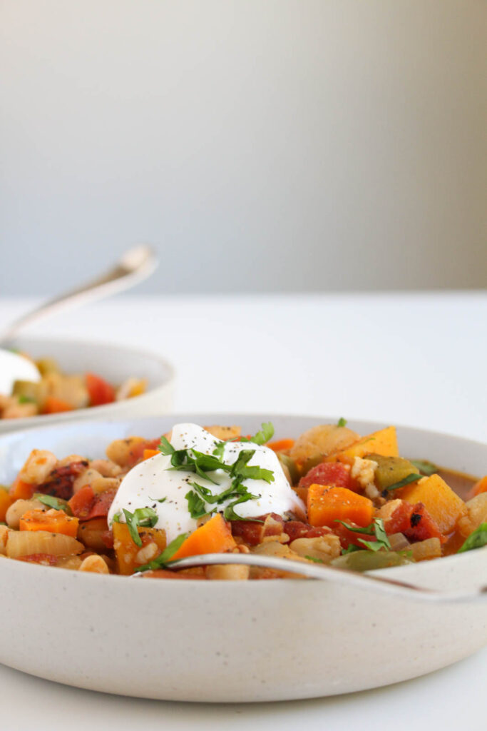 side view of butternut squash chili, with sour cream and herbs on top, in a ceramic bowl and spoon sitting in the middle